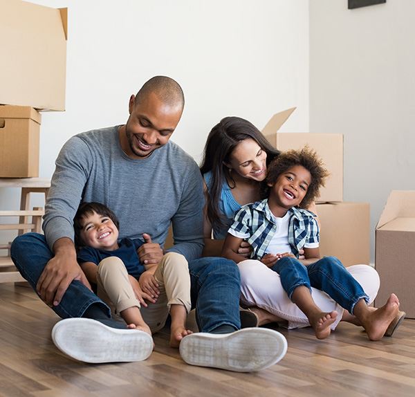 Mom, dad and two young boys smiling and sitting on a floor surrounded by moving boxes