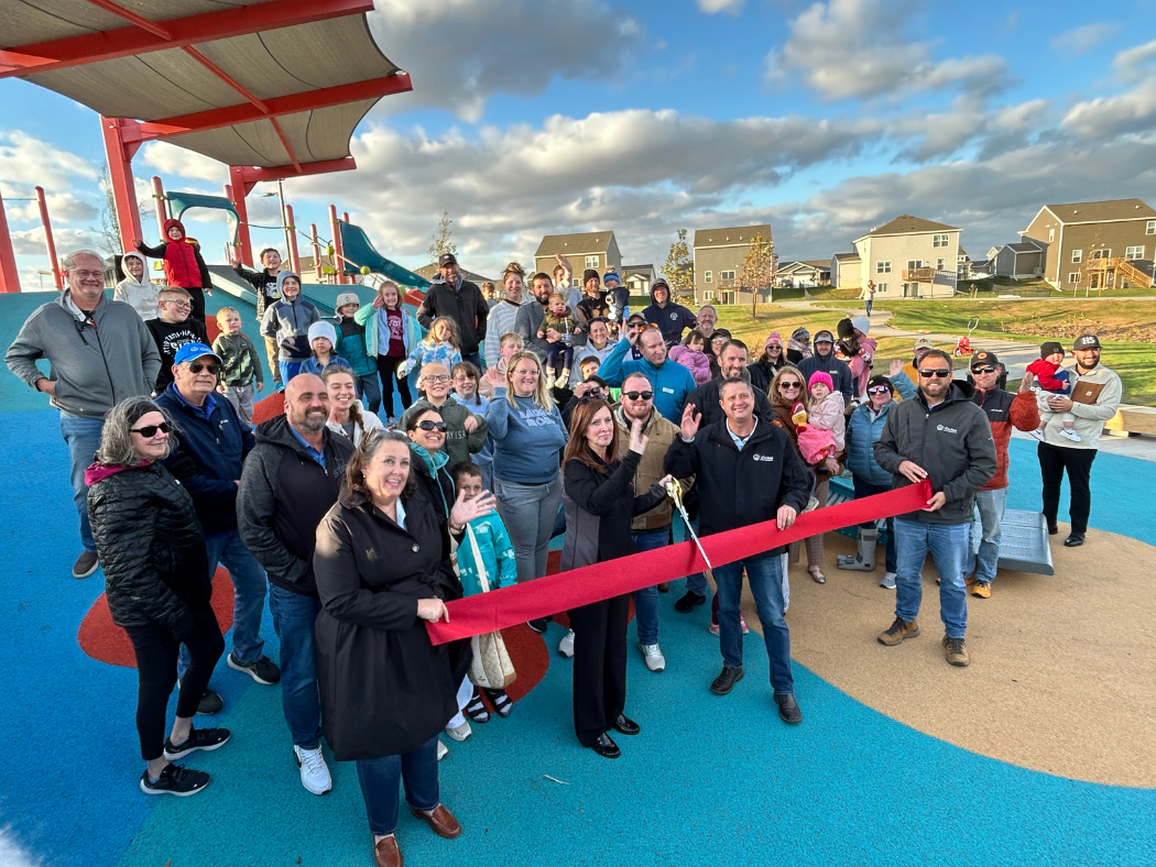 group of 50 smiling people gathered on playground to cut ribbon at park opening