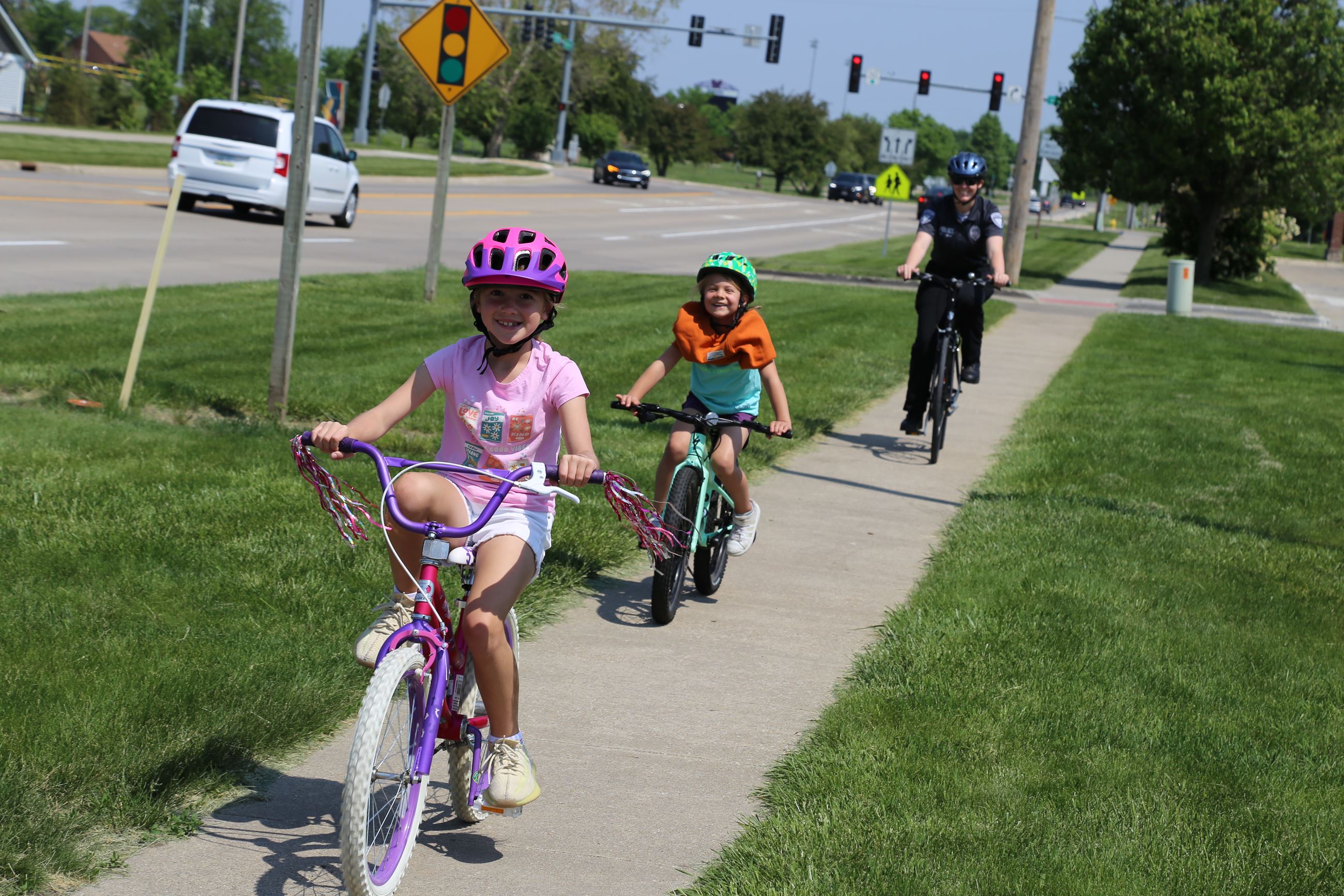 Two students joyfully ride their colorful bikes followed by a police officer smiling on a bicycle.